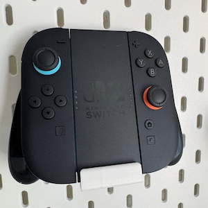 Op de afbeelding: Zwarte Nintendo Switch-controller met blauwe en oranje accenten, gemonteerd op een witte pegboard. De controller heeft knoppen, richtingspads en het Nintendo Switch-logo.