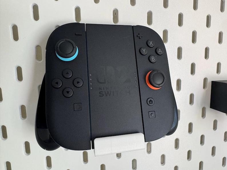 Op de afbeelding: Zwarte Nintendo Switch-controller met blauwe en oranje accenten, gemonteerd op een witte pegboard. De controller heeft knoppen, richtingspads en het Nintendo Switch-logo.