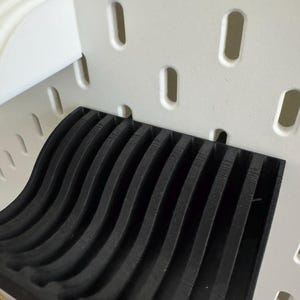 以下が含まれることがあります： 白のペグボードシステムに適合するように設計された、黒色の波模様の棚インサート。インサートには一連の垂直な隆起があり、テクスチャード加工された表面を作り出しています。ペグボードには楕円形の穴があります。