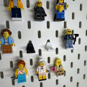 K&ouml;nnte beinhalten: Eine Sammlung von Lego-Minifiguren, jede mit einzigartigen Outfits und Accessoires. Zu den Figuren geh&ouml;ren ein Polizist, ein Koch und eine Frau, die eine Babyflasche h&auml;lt. Sie werden auf einer wei&szlig;en Lochplatte ausgestellt.