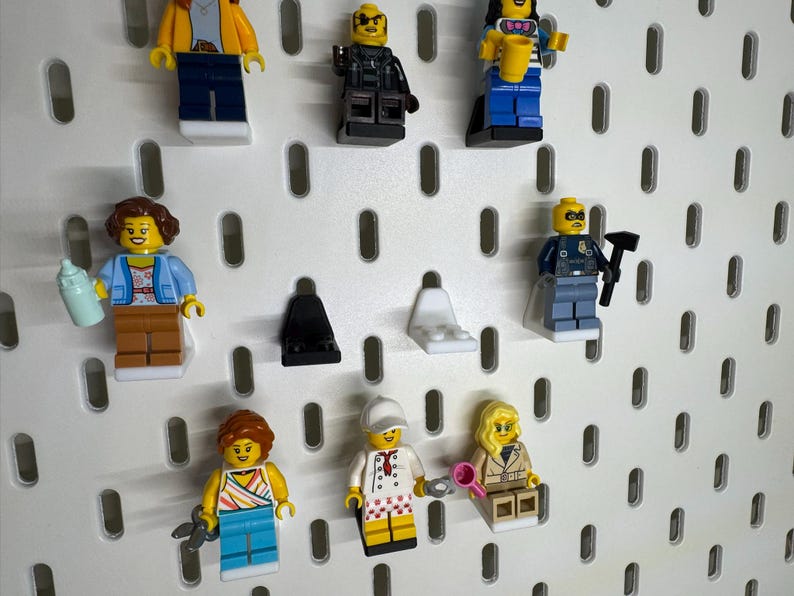 K&ouml;nnte beinhalten: Eine Sammlung von Lego-Minifiguren, jede mit einzigartigen Outfits und Accessoires. Zu den Figuren geh&ouml;ren ein Polizist, ein Koch und eine Frau, die eine Babyflasche h&auml;lt. Sie werden auf einer wei&szlig;en Lochplatte ausgestellt.