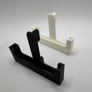 Op de afbeelding: Twee 3D-geprinte telefoonstandaards. E&eacute;n is zwart, de andere is wit. Elke standaard heeft een rechthoekige basis met een verticale steun en een kleine richel om een telefoon vast te houden. De standaards staan op een witte achtergrond.