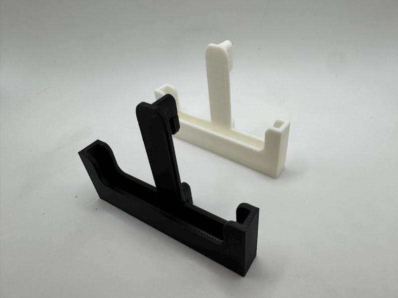 Op de afbeelding: Twee 3D-geprinte telefoonstandaards. E&eacute;n is zwart, de andere is wit. Elke standaard heeft een rechthoekige basis met een verticale steun en een kleine richel om een telefoon vast te houden. De standaards staan op een witte achtergrond.