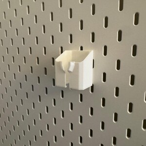May include: A white, 3D-printed storage cup attached to a pegboard. The cup has a rectangular shape with an open top and a small, angled section on the side. The pegboard is light gray with evenly spaced, oval-shaped holes.