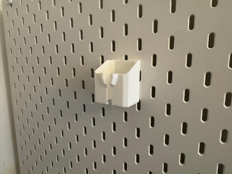 May include: A white, 3D-printed storage cup attached to a pegboard. The cup has a rectangular shape with an open top and a small, angled section on the side. The pegboard is light gray with evenly spaced, oval-shaped holes.