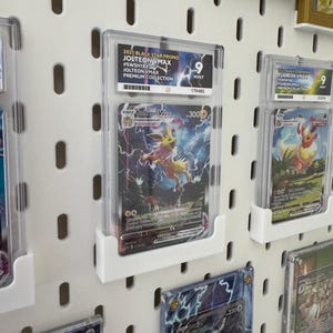 Może przedstawiać: Biała tablica z otworami z kilkoma kartami kolekcjonerskimi Pokemon w ochronnych etui. Karty są wyświetlane w rzędach i kolumnach. Wszystkie karty są ocenione i mają ocenę stanu idealnego. Wszystkie karty pochodzą z zestawu Black Star Promo z 2021 roku. Karty obejmują Jolteon VMAX i Flareon VMAX.