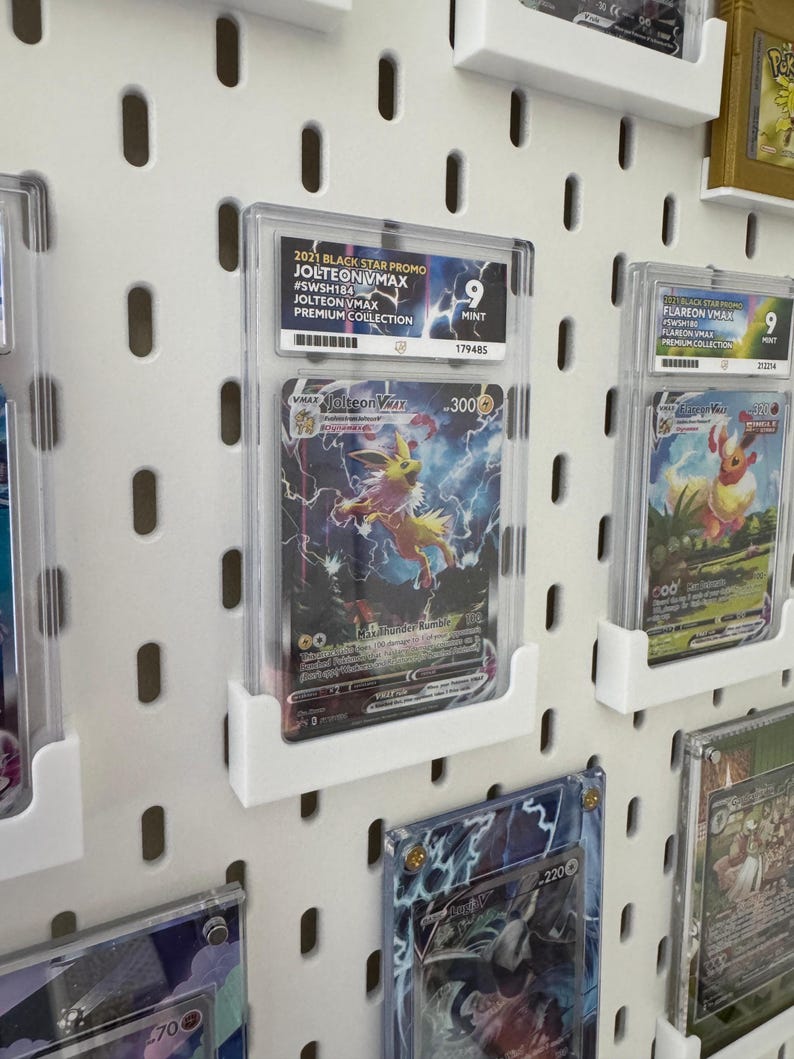 Może przedstawiać: Biała tablica z otworami z kilkoma kartami kolekcjonerskimi Pokemon w ochronnych etui. Karty są wyświetlane w rzędach i kolumnach. Wszystkie karty są ocenione i mają ocenę stanu idealnego. Wszystkie karty pochodzą z zestawu Black Star Promo z 2021 roku. Karty obejmują Jolteon VMAX i Flareon VMAX.