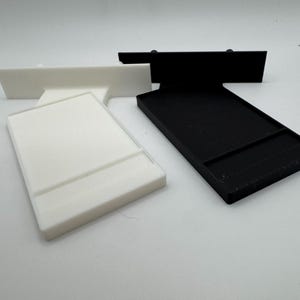 Puede incluir: Dos soportes para tel&eacute;fono impresos en 3D, uno blanco y otro negro. Ambos soportes tienen un borde elevado para sujetar un tel&eacute;fono.