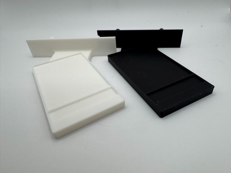 Puede incluir: Dos soportes para tel&eacute;fono impresos en 3D, uno blanco y otro negro. Ambos soportes tienen un borde elevado para sujetar un tel&eacute;fono.