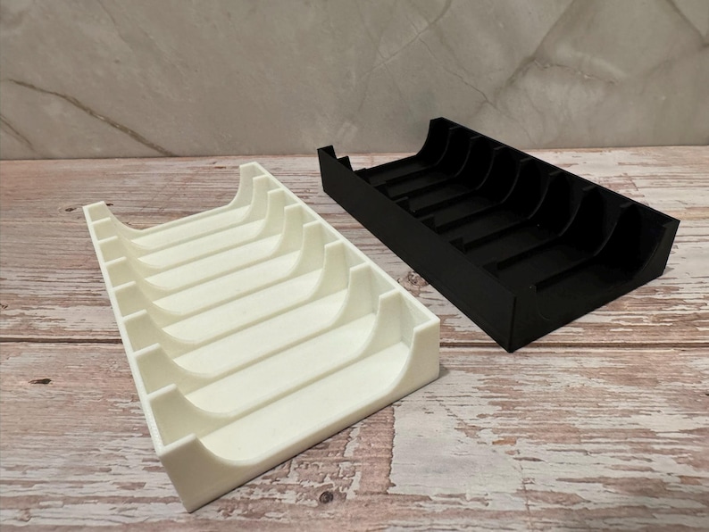 K&ouml;nnte beinhalten: Zwei rechteckige Organizer, einer wei&szlig; und einer schwarz, nebeneinander. Jeder Organizer hat gebogene Trennw&auml;nde, die einzelne F&auml;cher bilden. Die Organizer bestehen aus einem glatten Material und stehen auf einer Holzoberfl&auml;che.