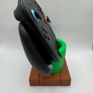 May include: Black video game controller on a brown and green stand. The stand is designed to resemble a green pipe on a brown brick base. The controller features blue and orange accents.