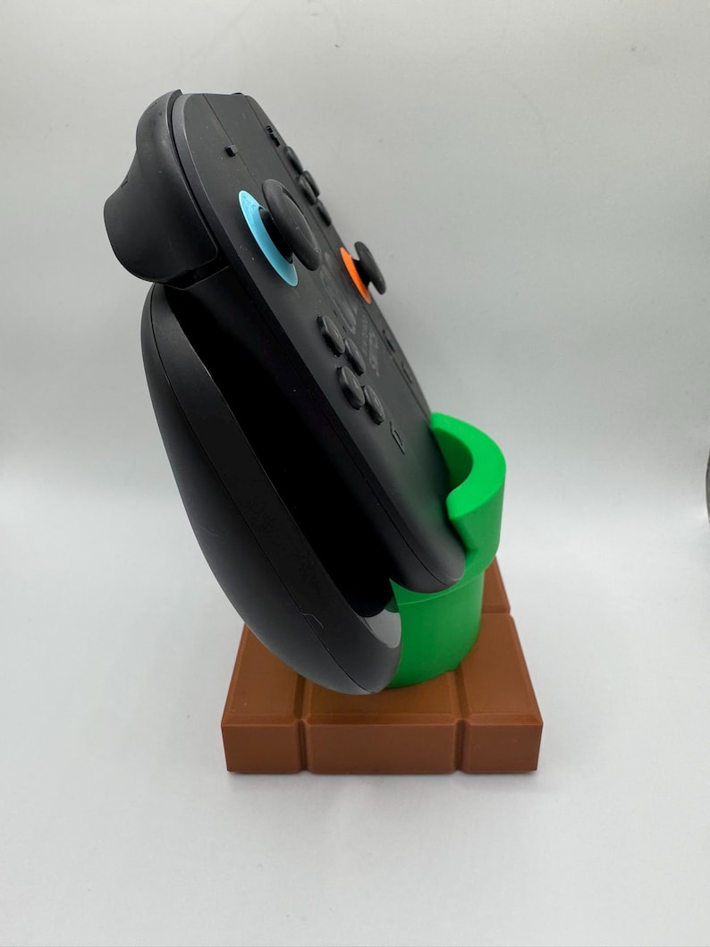 May include: Black video game controller on a brown and green stand. The stand is designed to resemble a green pipe on a brown brick base. The controller features blue and orange accents.