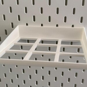 Op de afbeelding: Wit plasticen plank met een rasterpatroon, ontworpen om aan een pegboardwand te hangen.