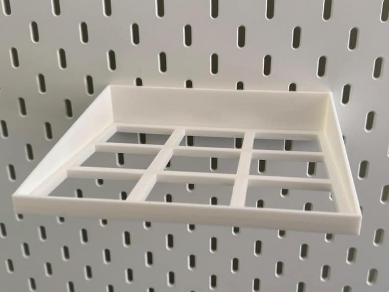 Op de afbeelding: Wit plasticen plank met een rasterpatroon, ontworpen om aan een pegboardwand te hangen.