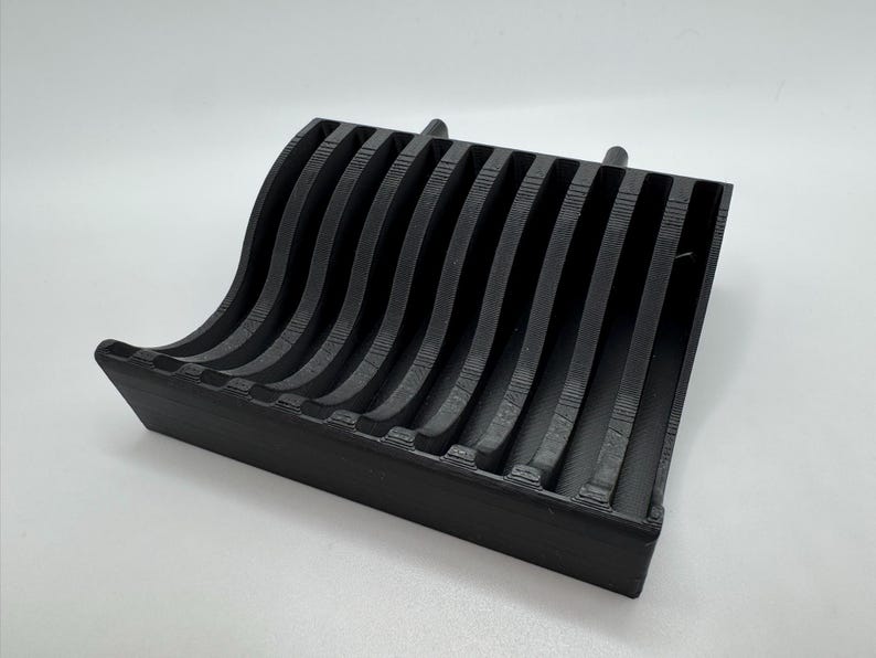 以下が含まれることがあります： 黒色の3Dプリントされた電話スタンド。波のような曲線的なデザインで、複数の垂直スロットがあり、デバイスを保持します。長方形のベースがあり、マットブラック素材で作られています。