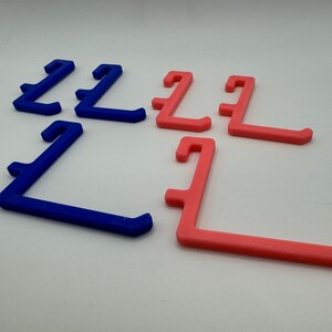 May include: A set of six plastic hooks, three blue and three pink, designed for hanging items. The hooks are shaped like an L and have a rounded edge.