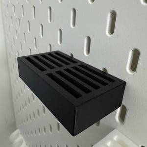 Op de afbeelding: Een zwarte rechthoekige pegboard plank met een sleufvormige bovenkant. De plank is gemonteerd op een witte pegboard met gelijkmatig verdeelde ovale gaten. De plank is ontworpen om kleine items op te bergen.