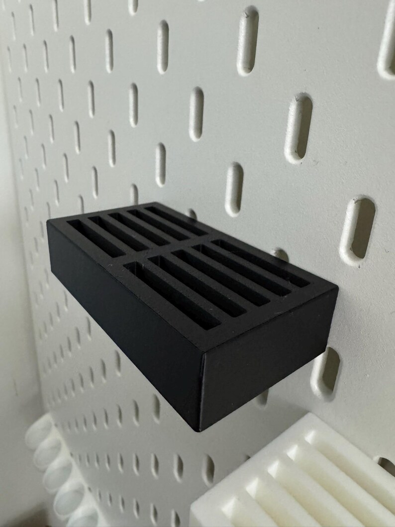 Op de afbeelding: Een zwarte rechthoekige pegboard plank met een sleufvormige bovenkant. De plank is gemonteerd op een witte pegboard met gelijkmatig verdeelde ovale gaten. De plank is ontworpen om kleine items op te bergen.