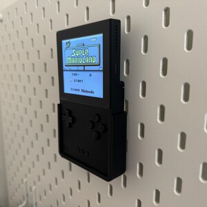 Op de afbeelding: Een zwarte draagbare videogameconsole met een scherm waarop het spel "Super Marioland" wordt weergegeven. De console is gemonteerd op een witte pegboard-wand.