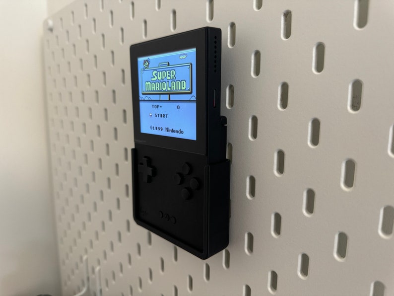 Op de afbeelding: Een zwarte draagbare videogameconsole met een scherm waarop het spel "Super Marioland" wordt weergegeven. De console is gemonteerd op een witte pegboard-wand.