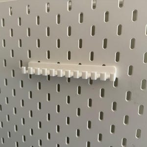 May include: A white pegboard with evenly spaced, oblong holes. A white pegboard accessory, designed to hold items, is attached to the board. The accessory has a series of small, vertical teeth. The pegboard and accessory are made of a smooth, solid material.