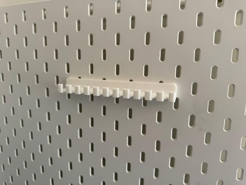 May include: A white pegboard with evenly spaced, oblong holes. A white pegboard accessory, designed to hold items, is attached to the board. The accessory has a series of small, vertical teeth. The pegboard and accessory are made of a smooth, solid material.
