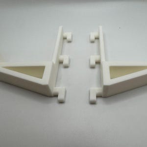 May include: Two white, angled support brackets. Each bracket has a triangular opening and a series of small, rectangular protrusions. The brackets are designed to provide support and stability.