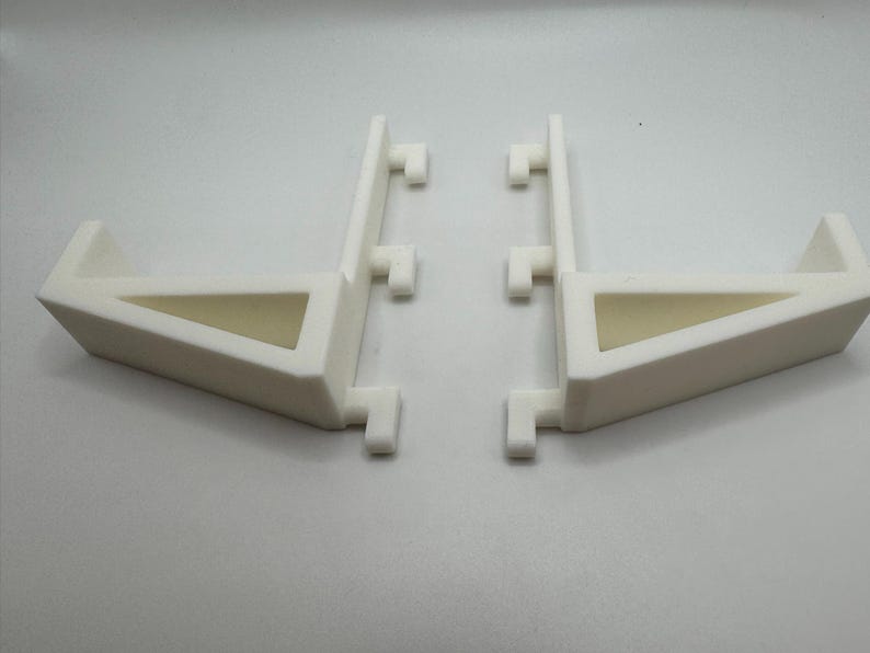 May include: Two white, angled support brackets. Each bracket has a triangular opening and a series of small, rectangular protrusions. The brackets are designed to provide support and stability.