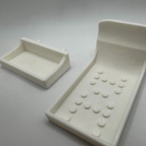 May include: Two white 3D-printed objects. One is a rectangular tray with a curved handle. The other is a larger, angled piece with a curved top and several circular indentations, resembling a miniature pinball machine.
