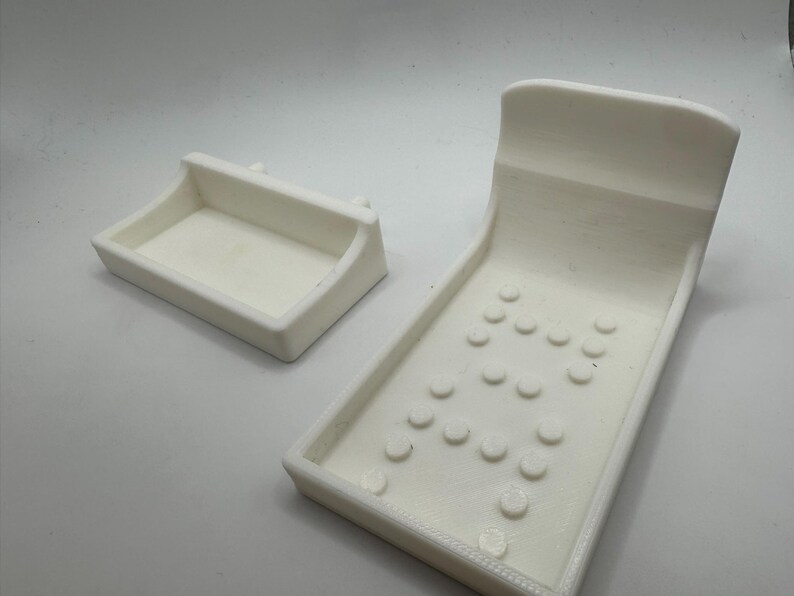 May include: Two white 3D-printed objects. One is a rectangular tray with a curved handle. The other is a larger, angled piece with a curved top and several circular indentations, resembling a miniature pinball machine.