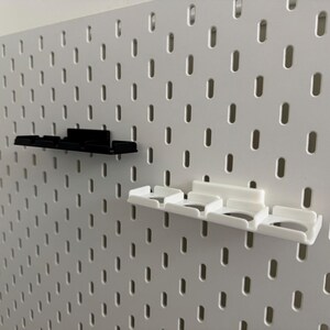 K&ouml;nnte beinhalten: Eine wei&szlig;e Lochwand mit zwei angebrachten Regalen. Ein Regal ist schwarz, das andere wei&szlig;. Die Regale haben eine rechteckige Form mit mehreren F&auml;chern. Die Lochwand hat viele gleichm&auml;&szlig;ig verteilte ovale L&ouml;cher.