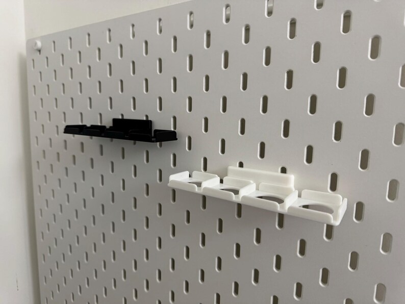 K&ouml;nnte beinhalten: Eine wei&szlig;e Lochwand mit zwei angebrachten Regalen. Ein Regal ist schwarz, das andere wei&szlig;. Die Regale haben eine rechteckige Form mit mehreren F&auml;chern. Die Lochwand hat viele gleichm&auml;&szlig;ig verteilte ovale L&ouml;cher.
