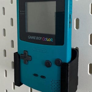 May include: A teal Game Boy Color handheld video game console with a black screen and buttons. The console is mounted on a black plastic wall mount.