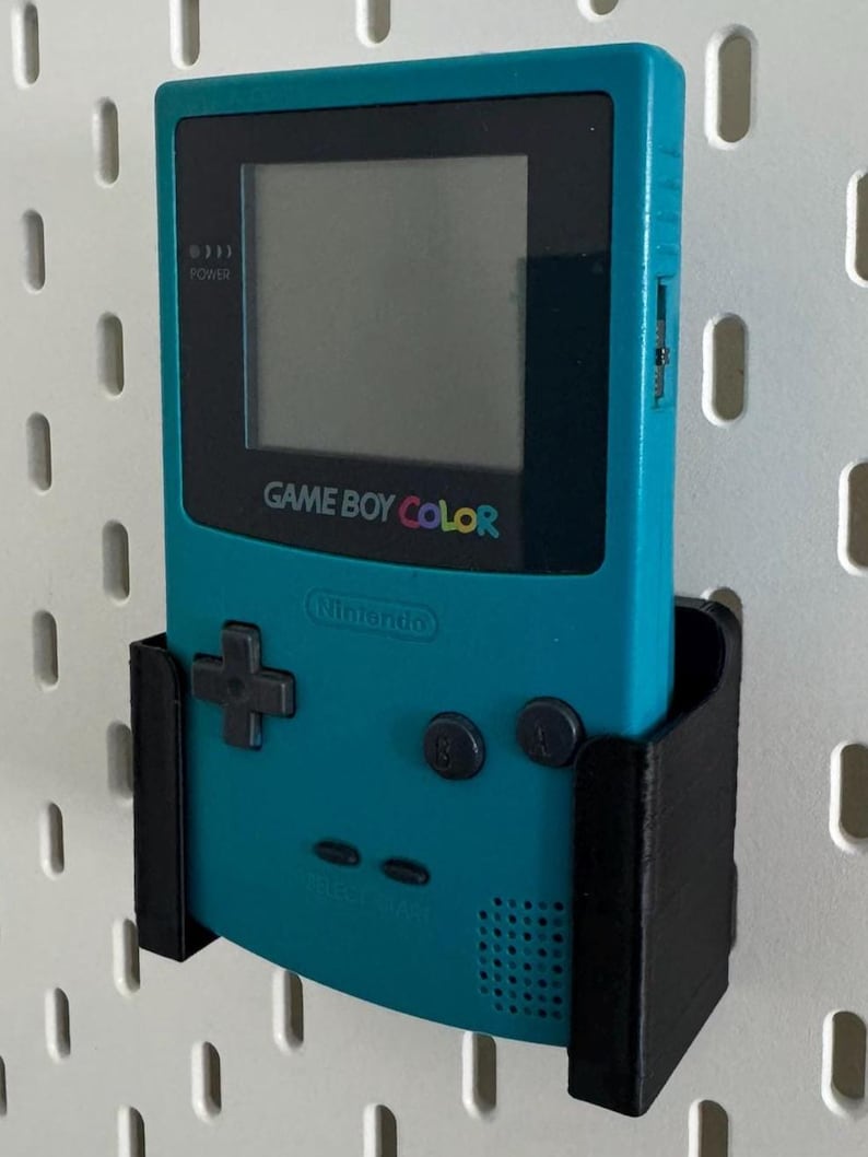 May include: A teal Game Boy Color handheld video game console with a black screen and buttons. The console is mounted on a black plastic wall mount.