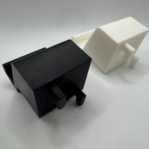 Op de afbeelding: Twee geometrische, rechthoekige objecten. De ene is zwart, de andere wit. Beide hebben een vergelijkbaar ontwerp met een rechthoekig lichaam en twee kleine, cilindrische uitsteeksels. De objecten zijn waarschijnlijk 3D-geprint en liggen op een wit oppervlak.