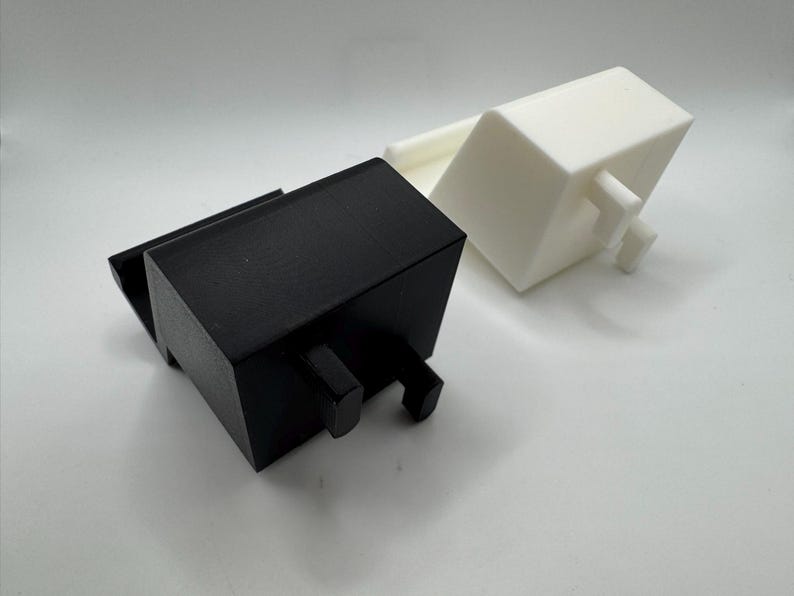 Op de afbeelding: Twee geometrische, rechthoekige objecten. De ene is zwart, de andere wit. Beide hebben een vergelijkbaar ontwerp met een rechthoekig lichaam en twee kleine, cilindrische uitsteeksels. De objecten zijn waarschijnlijk 3D-geprint en liggen op een wit oppervlak.