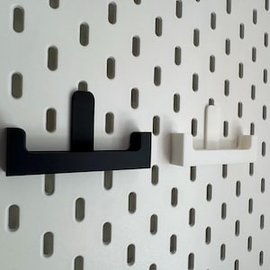 May include: Two wall-mounted storage shelves, one black and one white, are displayed against a white pegboard background. The shelves have a simple, modern design with a flat base and a raised back. The pegboard has evenly spaced oval holes.