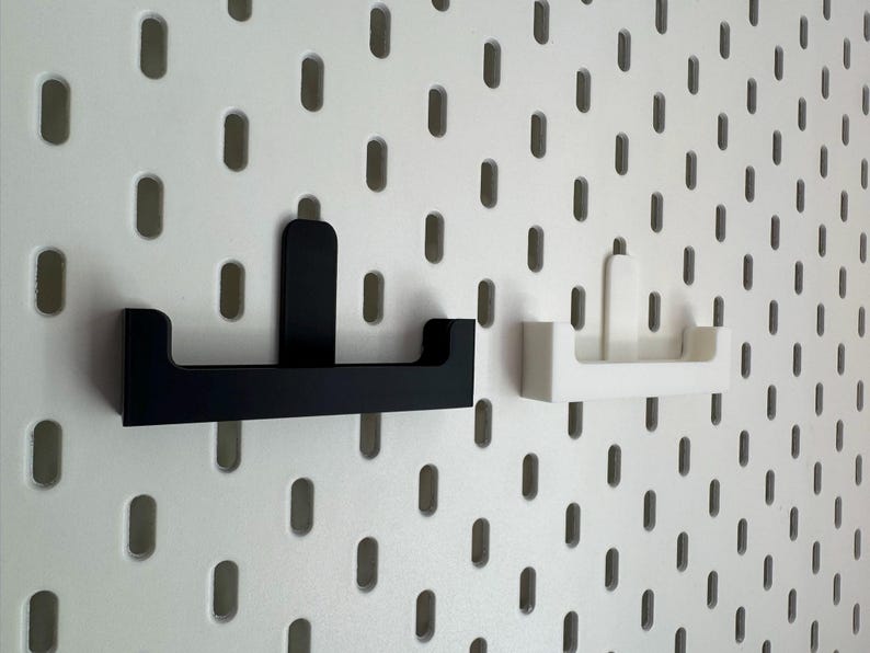May include: Two wall-mounted storage shelves, one black and one white, are displayed against a white pegboard background. The shelves have a simple, modern design with a flat base and a raised back. The pegboard has evenly spaced oval holes.