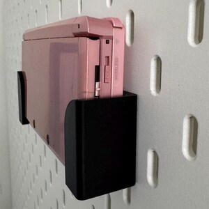 Pode incluir: Um console de jogos port&aacute;til Nintendo 3DS rosa com um suporte de parede preto. O console tem um r&oacute;tulo branco que diz "Wireless" no lado.