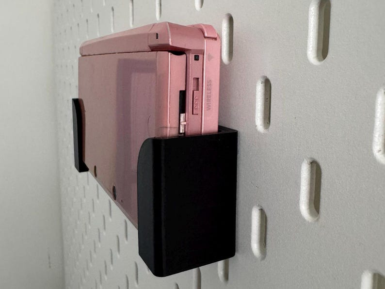 Pode incluir: Um console de jogos port&aacute;til Nintendo 3DS rosa com um suporte de parede preto. O console tem um r&oacute;tulo branco que diz "Wireless" no lado.