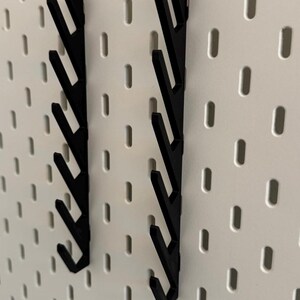 Pu&ograve; includere: Due ganci in plastica nera per pannello forato con un design a zig-zag. I ganci sono montati su un pannello forato bianco.