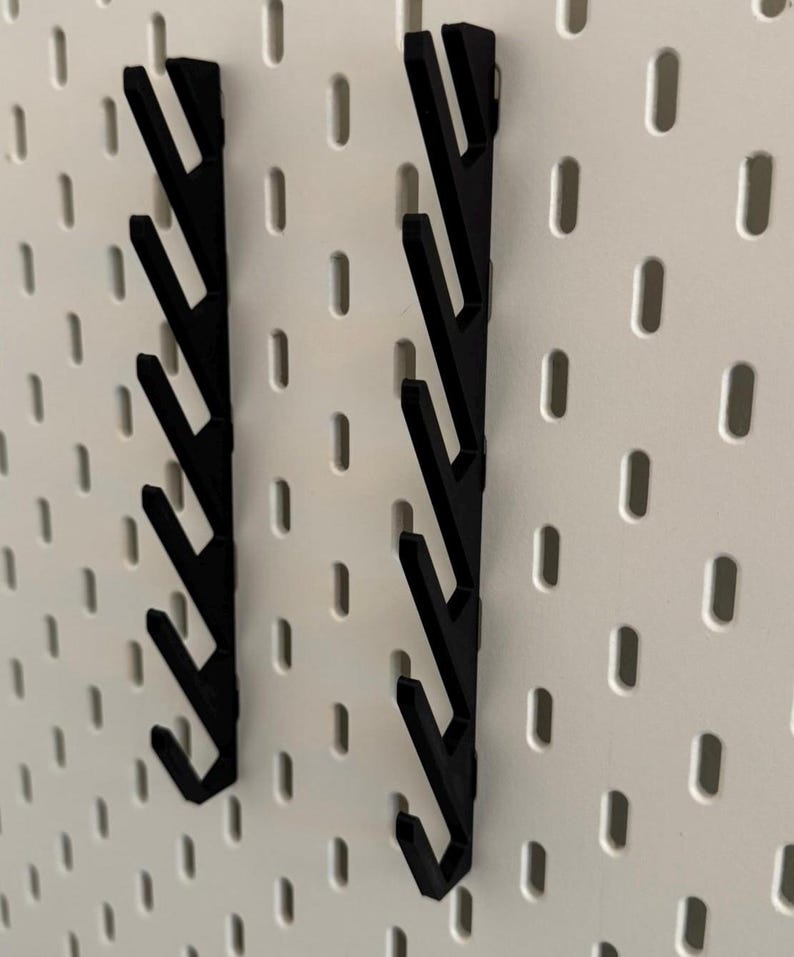Pu&ograve; includere: Due ganci in plastica nera per pannello forato con un design a zig-zag. I ganci sono montati su un pannello forato bianco.