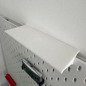 Puede incluir: Un tablero de clavijas blanco con una repisa blanca unida a la parte superior. Un dispositivo de juego port&aacute;til plateado est&aacute; colgado del tablero de clavijas.