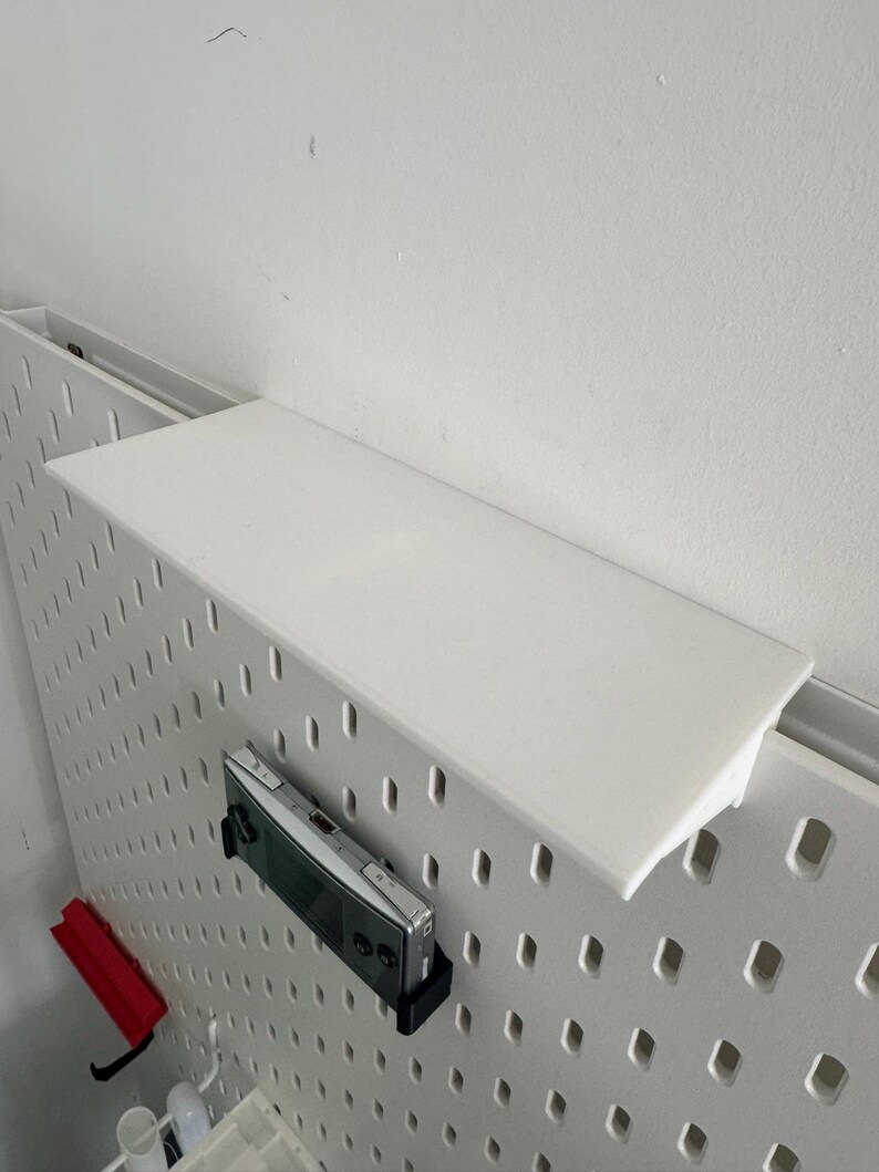 Puede incluir: Un tablero de clavijas blanco con una repisa blanca unida a la parte superior. Un dispositivo de juego port&aacute;til plateado est&aacute; colgado del tablero de clavijas.