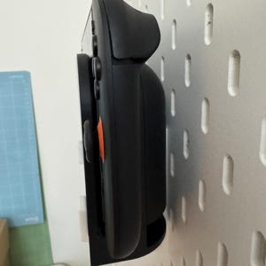 Op de afbeelding: Zwarte gamecontroller gemonteerd op een witte pegboard. De controller heeft een oranje accent en wordt vastgehouden door een zwarte beugel. De pegboard heeft gelijkmatig verdeelde gaten.