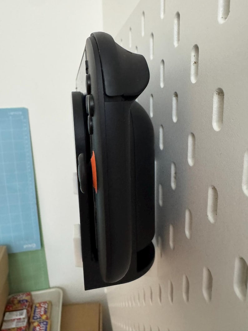 Op de afbeelding: Zwarte gamecontroller gemonteerd op een witte pegboard. De controller heeft een oranje accent en wordt vastgehouden door een zwarte beugel. De pegboard heeft gelijkmatig verdeelde gaten.