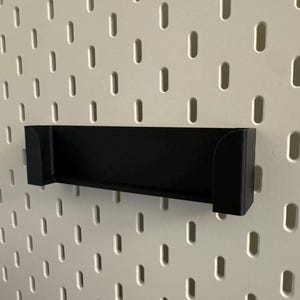 Puede incluir: Un estante negro para panel perforado, dise&ntilde;ado para fijarse a un panel perforado. El estante es rectangular con lados curvos y est&aacute; montado en un panel perforado blanco con agujeros ovalados espaciados.