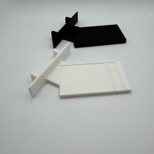 Puede incluir: Dos expositores de pl&aacute;stico, uno blanco y otro negro. Ambos expositores tienen una base rectangular y un peque&ntilde;o soporte en forma de T.