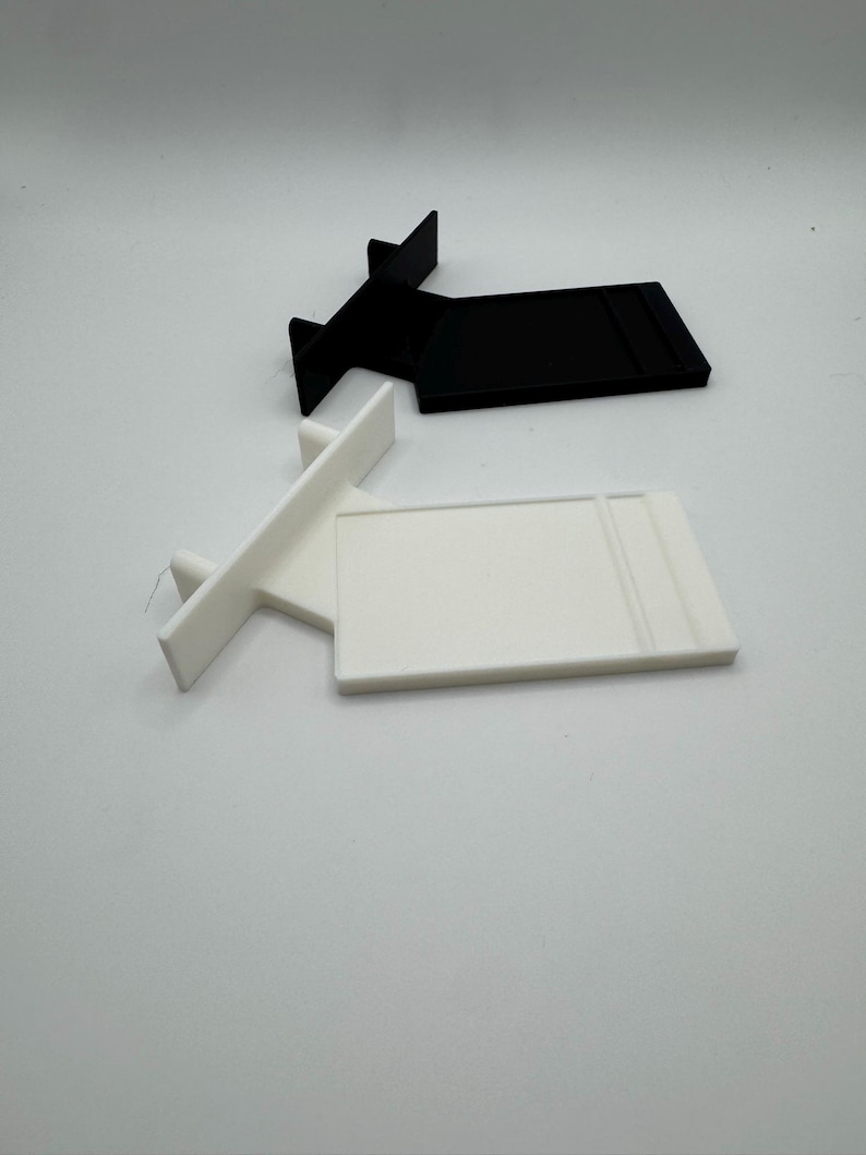 Puede incluir: Dos expositores de pl&aacute;stico, uno blanco y otro negro. Ambos expositores tienen una base rectangular y un peque&ntilde;o soporte en forma de T.