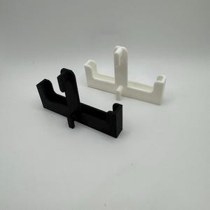 Puede incluir: Dos objetos geom&eacute;tricos de pl&aacute;stico, uno negro y otro blanco. Ambos tienen una base rectangular con una secci&oacute;n elevada y una proyecci&oacute;n en forma de gancho. Los objetos est&aacute;n dise&ntilde;ados para sujetar o soportar algo.